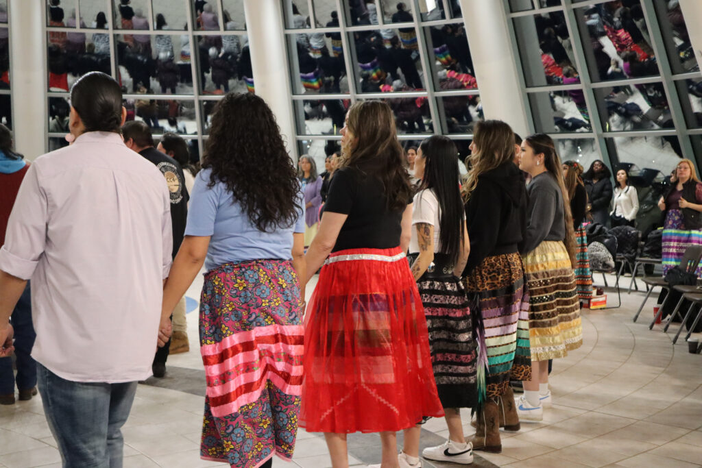 People holding hands in a circle, wearing traditional Indigenous clothing.
